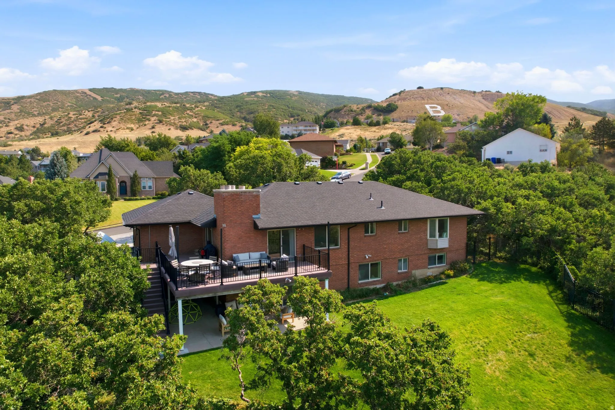 Brick exterior residence with a spacious elevated deck, tiered landscaping, expansive green lawn, and mature trees
