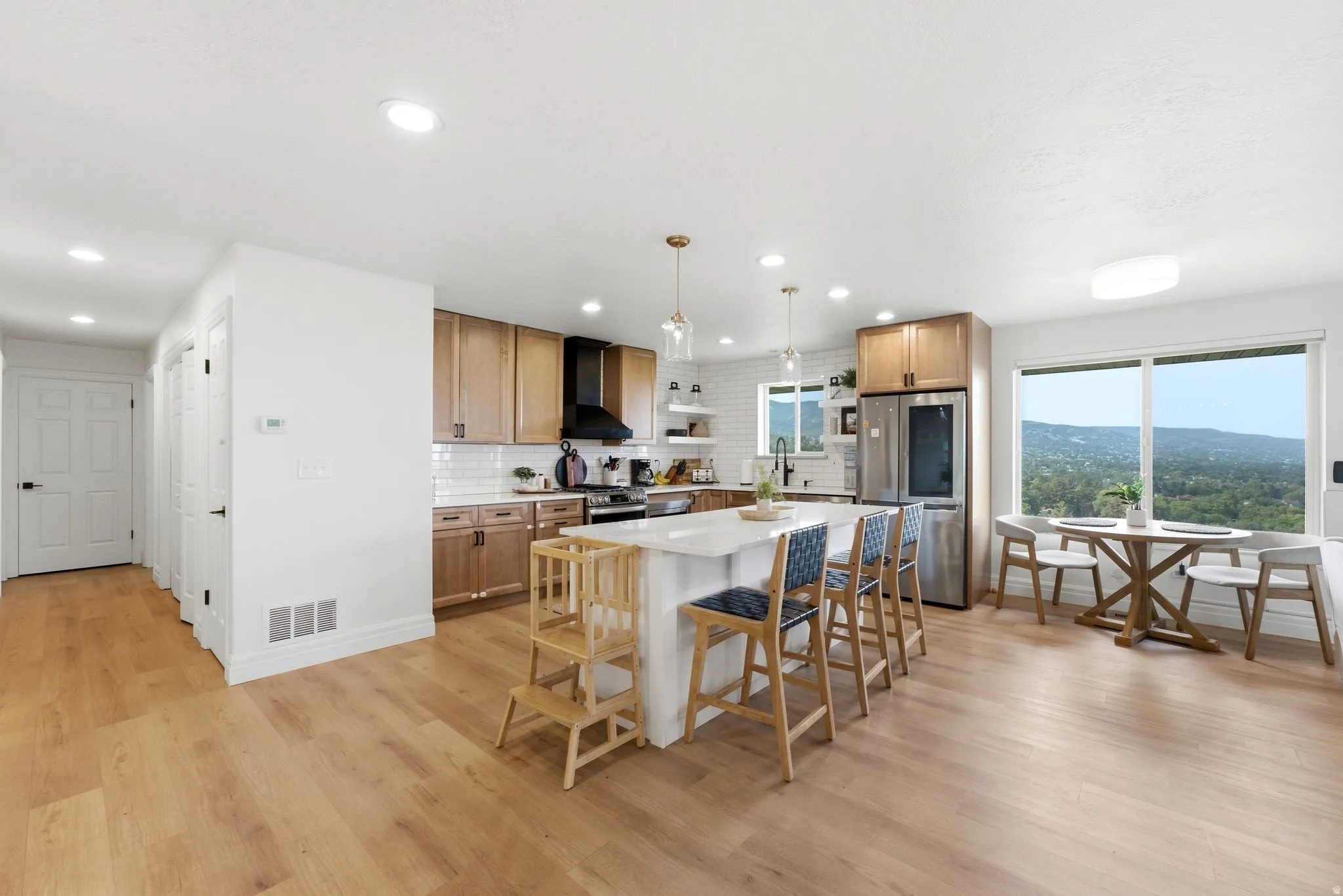 Open-concept kitchen featuring light wood-finish flooring, a central island with a light countertop, wood cabinetry, stainless steel appliances, and large windows revealing a distant landscape