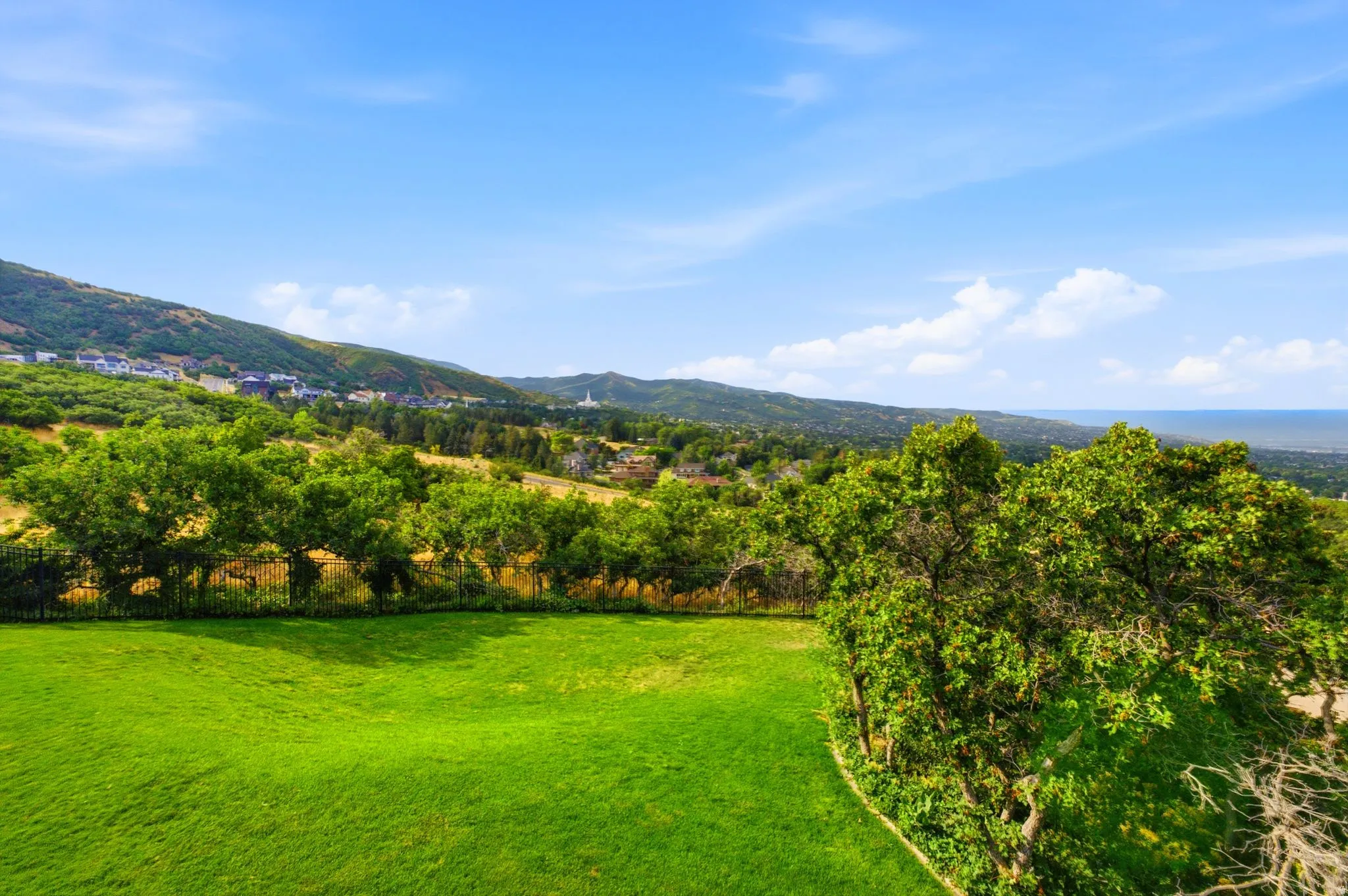 Expansive green lawn with mature trees and a black metal fence, complemented by distant hillside homes and a visible body of water