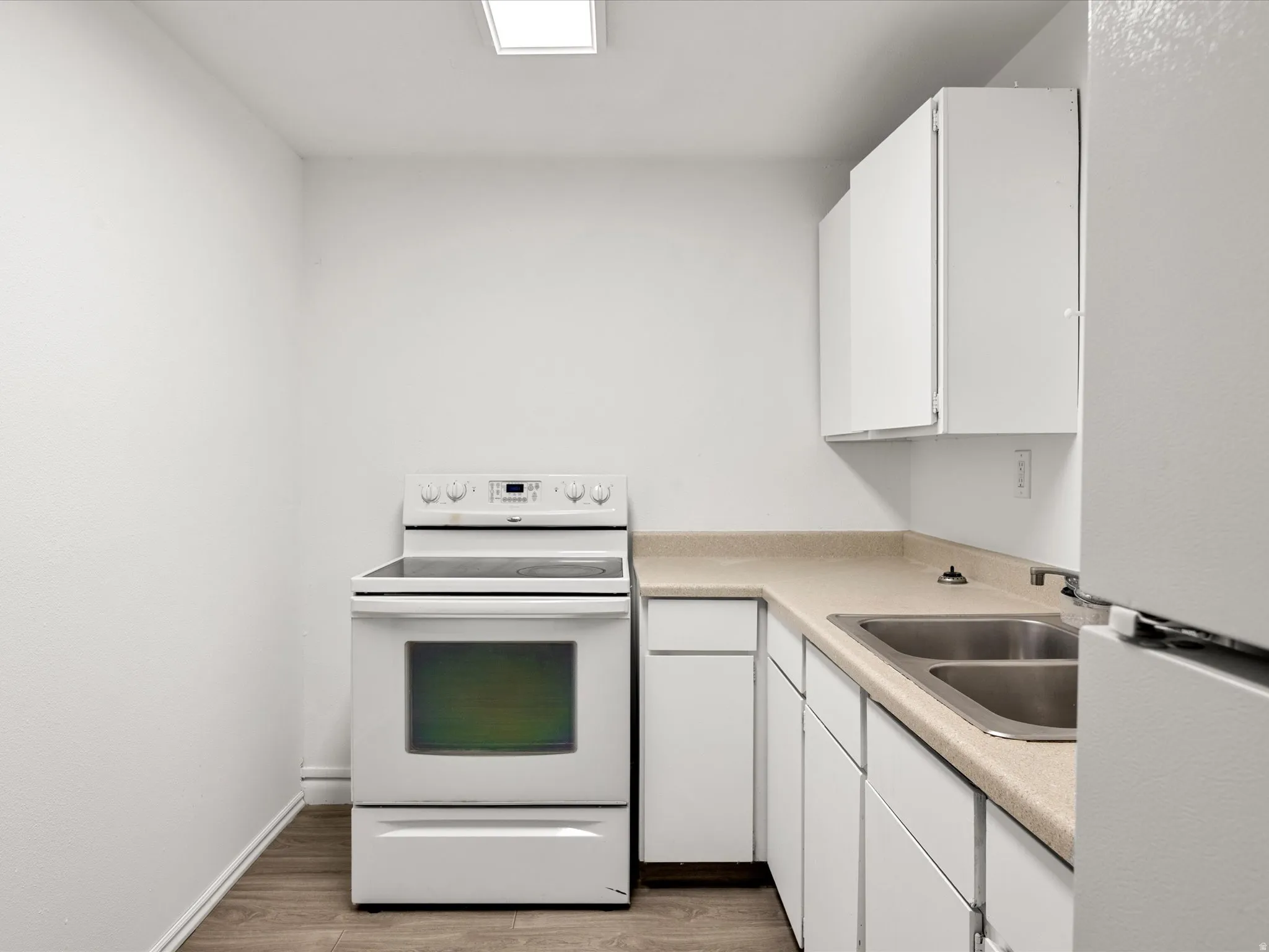 Compact kitchen featuring white cabinetry, a double basin stainless steel sink, light-toned countertops, a white electric range, and wood-finish flooring