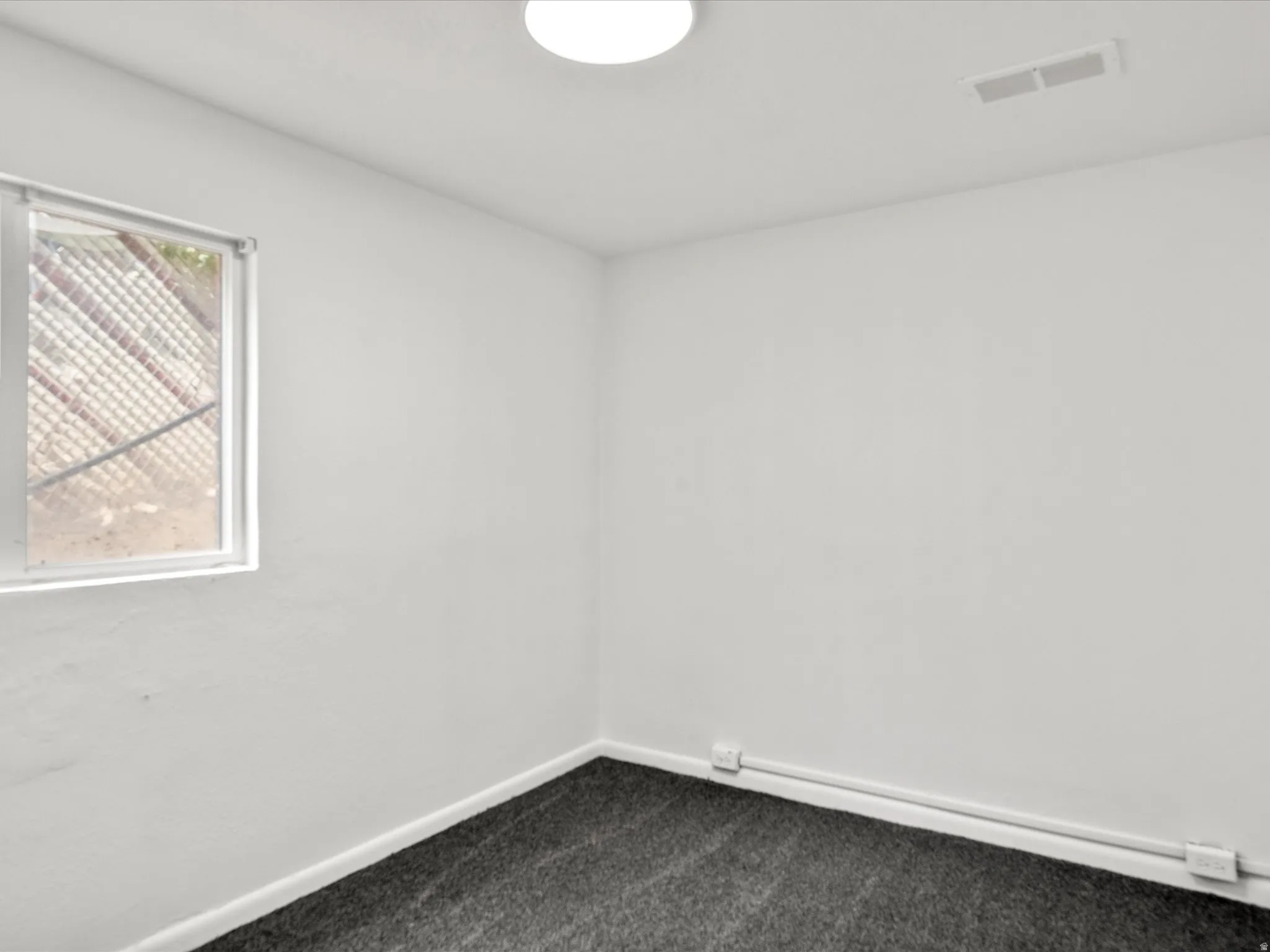 Neutral-toned walls and dark grey carpeting define this room, featuring a single window and an overhead flush-mount light fixture