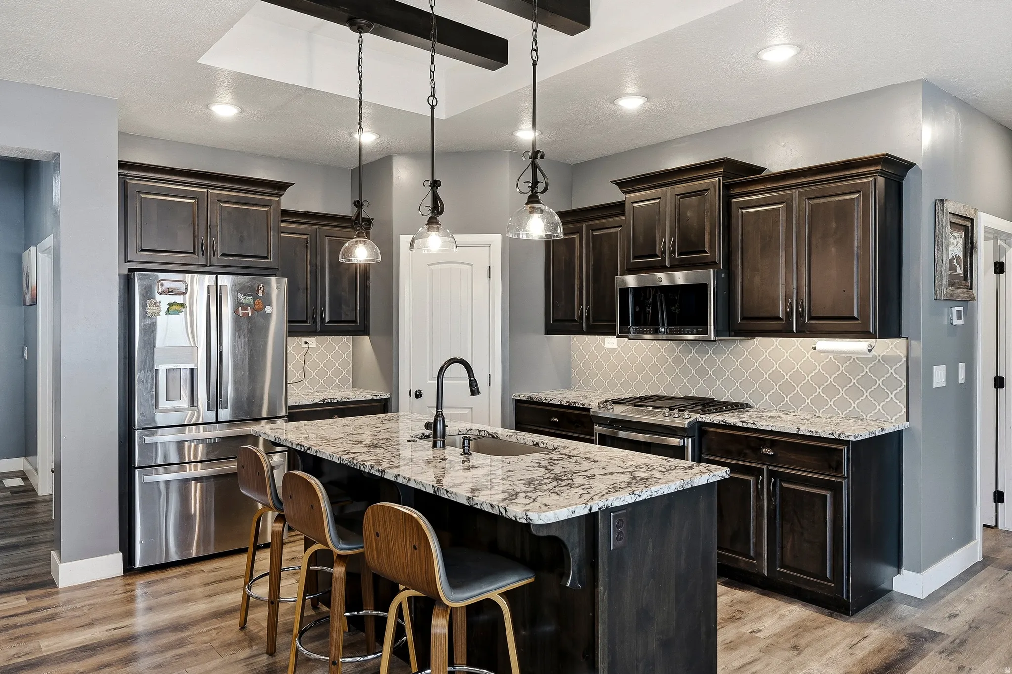 Spacious kitchen featuring an island with a built-in sink and pendant lighting, dark wood-finish cabinetry, stainless steel appliances, and a tiled backsplash