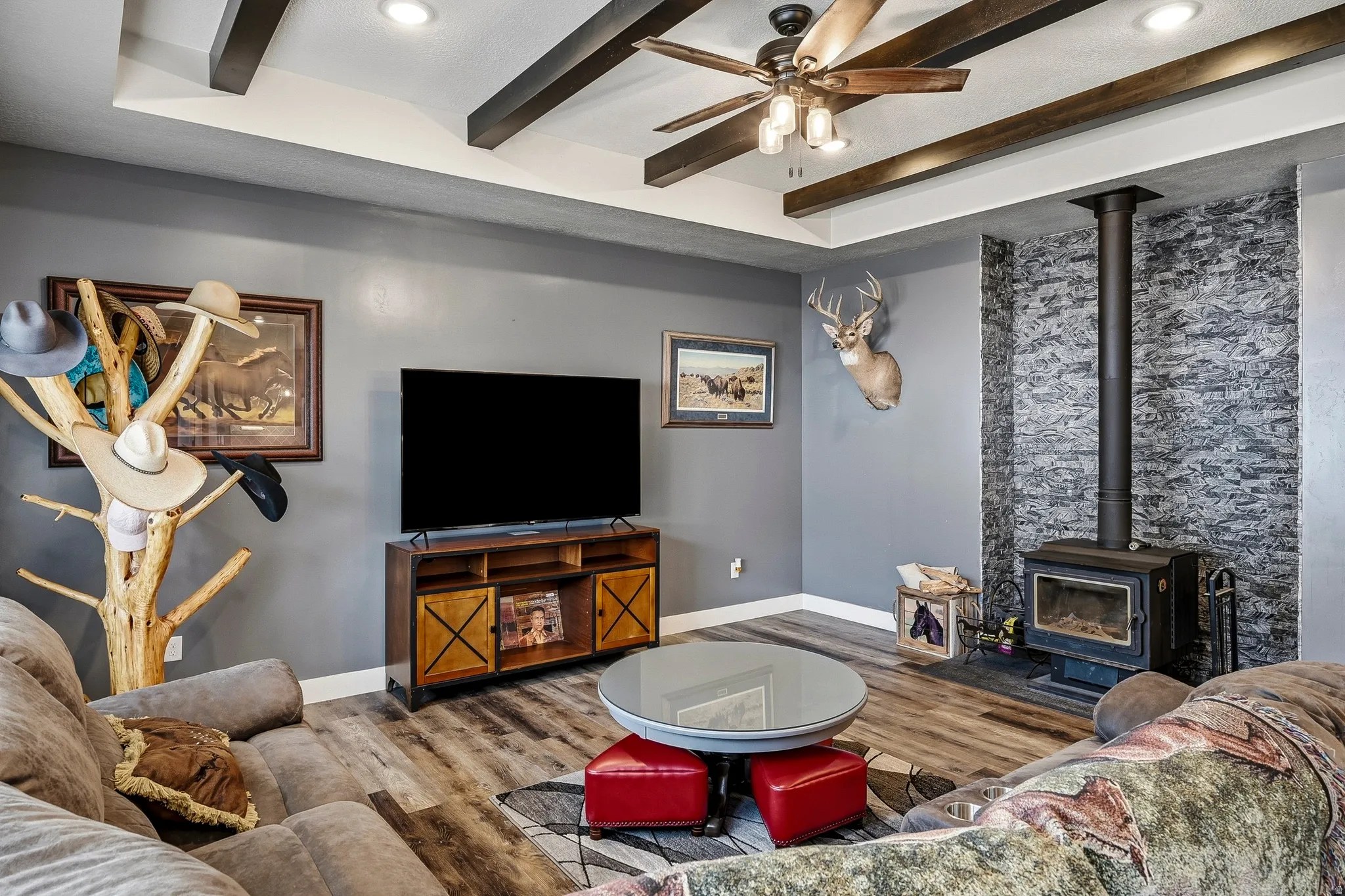 Living area featuring a stone veneer accent wall with a wood-burning stove, wood-finish flooring, a ceiling fan with light kit, and exposed wood ceiling beams