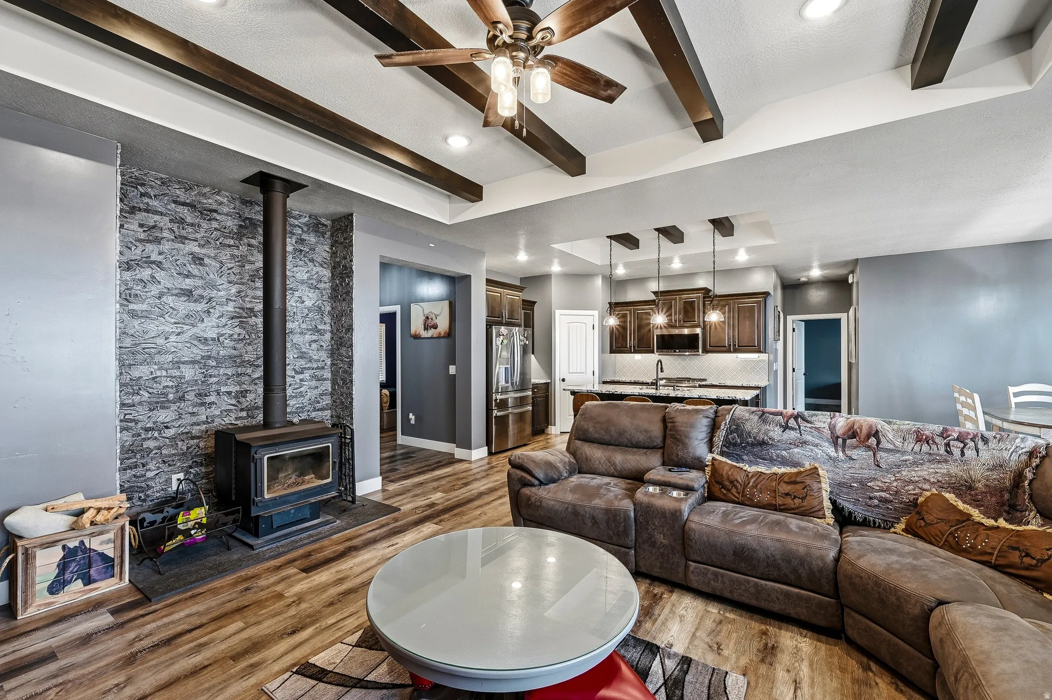 Living area featuring a stone accent wall with a black wood-burning stove, wood-finish flooring, recessed lighting, and exposed ceiling beams