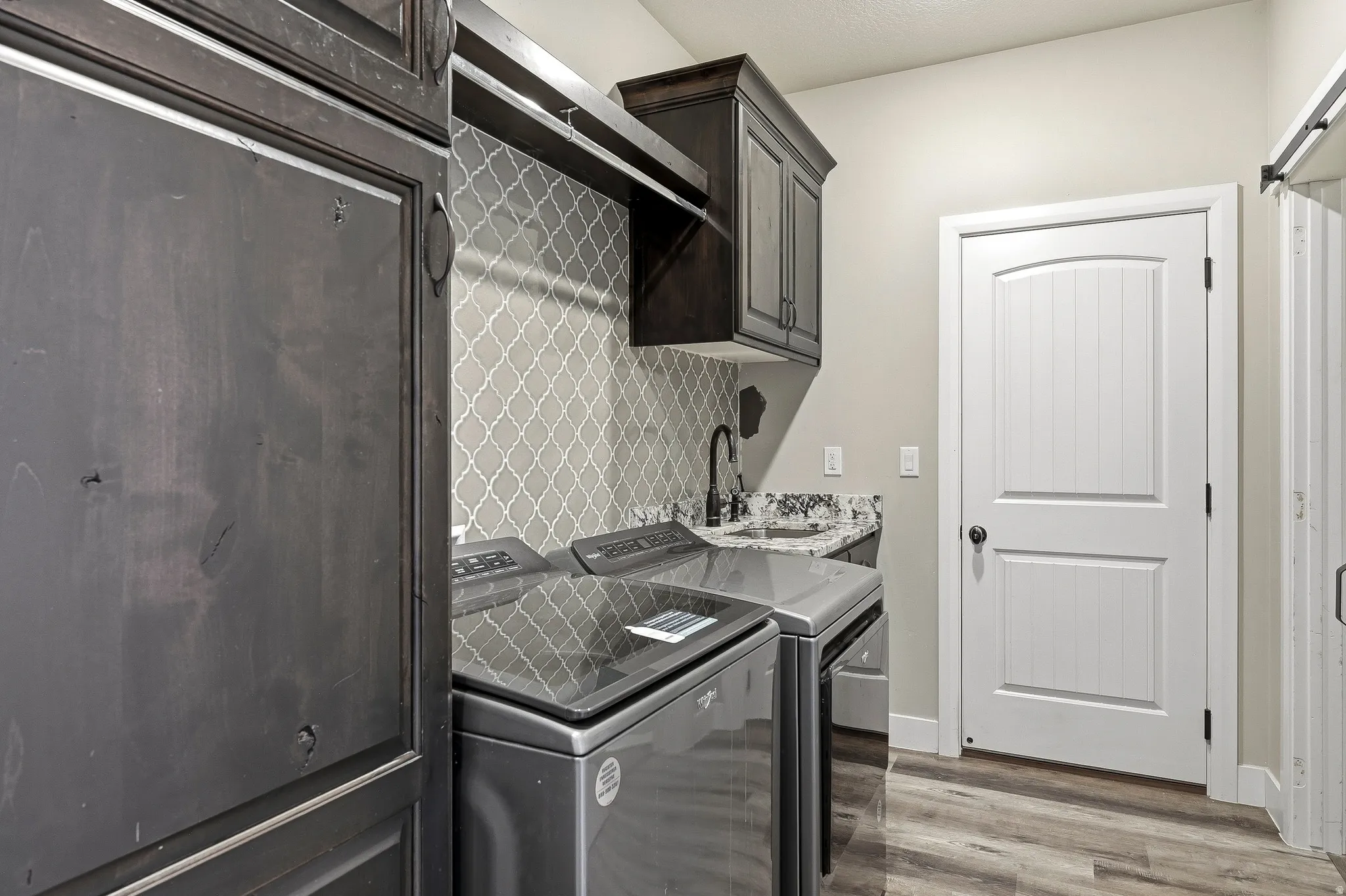 Laundry area featuring dark wood-finish cabinetry, a built-in sink with a black faucet, gray arabesque tile backsplash, and wood-finish flooring