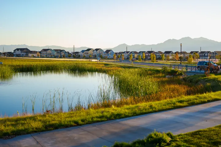 Community pond with natural landscaping, pedestrian path, residential street, and distant mountain backdrop