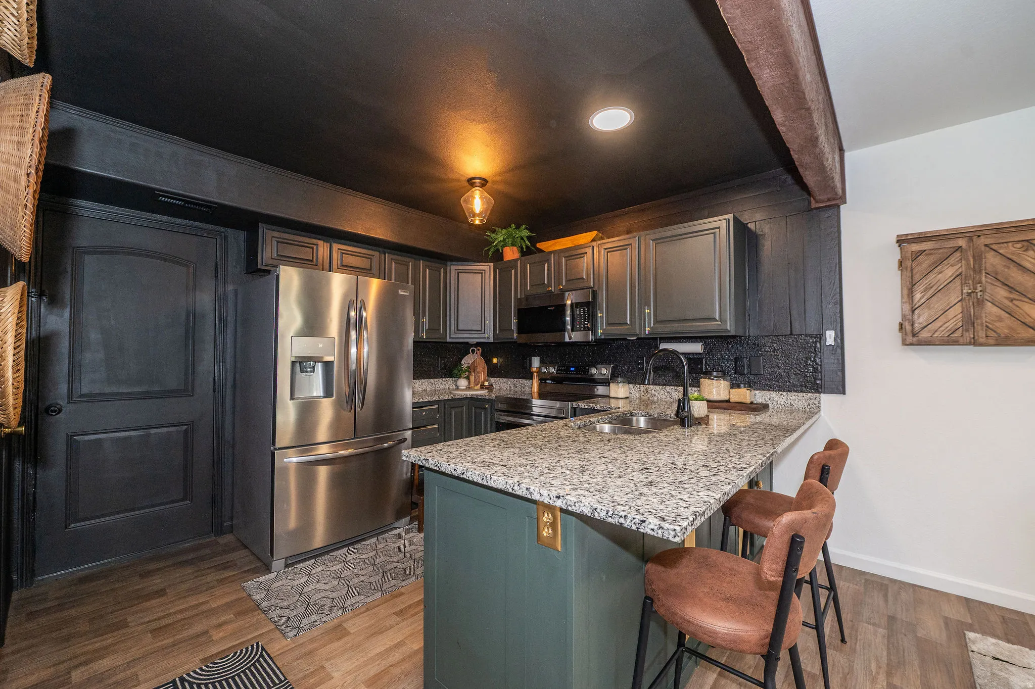 Modern kitchen featuring granite countertops, stainless steel appliances, dark cabinetry, wood-finish flooring, and a textured black backsplash