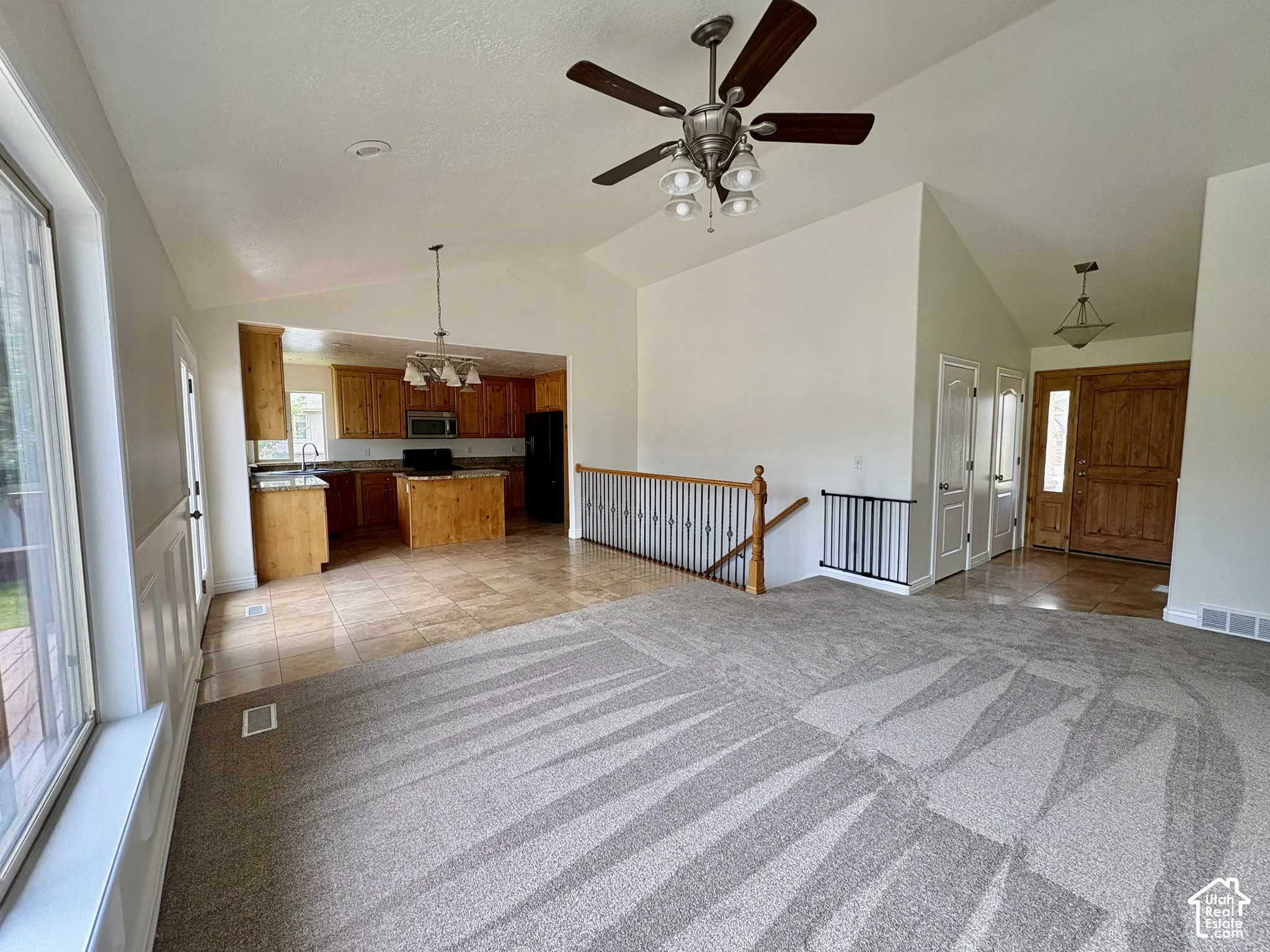 Vaulted great room with a ceiling fan, a kitchen featuring wood cabinetry and an island, a wood-finish front door with an adjacent window, and a carpeted main floor