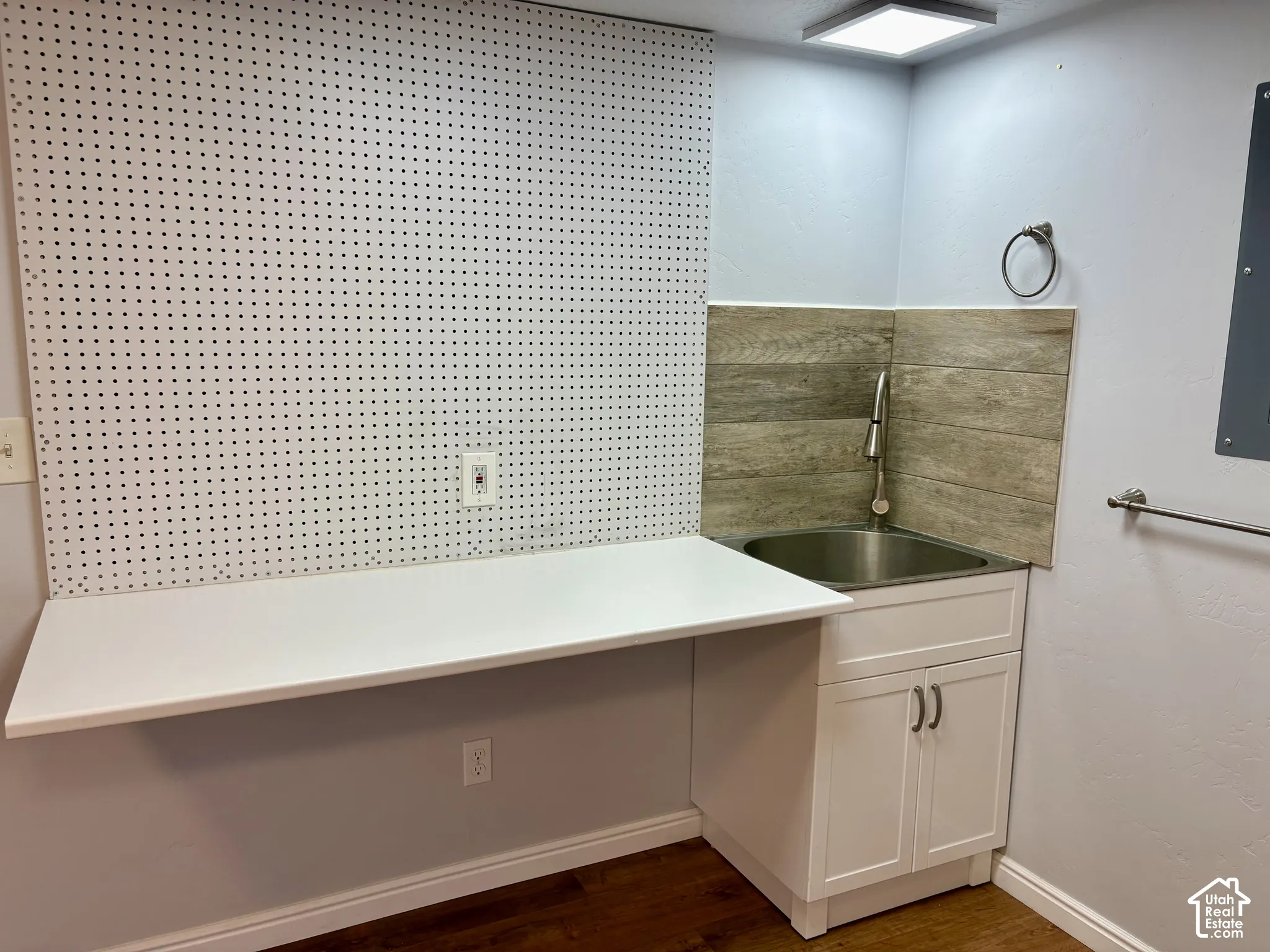 Utility area featuring a stainless steel sink, white shaker-style cabinetry, wood-finish flooring, a wall-mounted pegboard, and a square ceiling light fixture