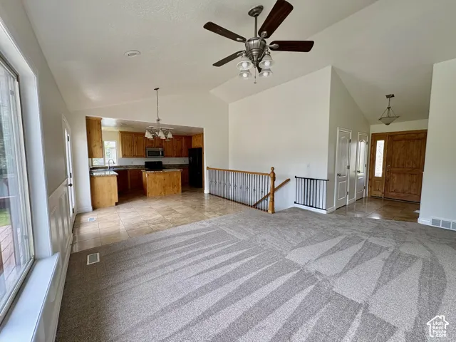 Vaulted great room with a ceiling fan, a kitchen featuring wood cabinetry and an island, a wood-finish front door with an adjacent window, and a carpeted main floor