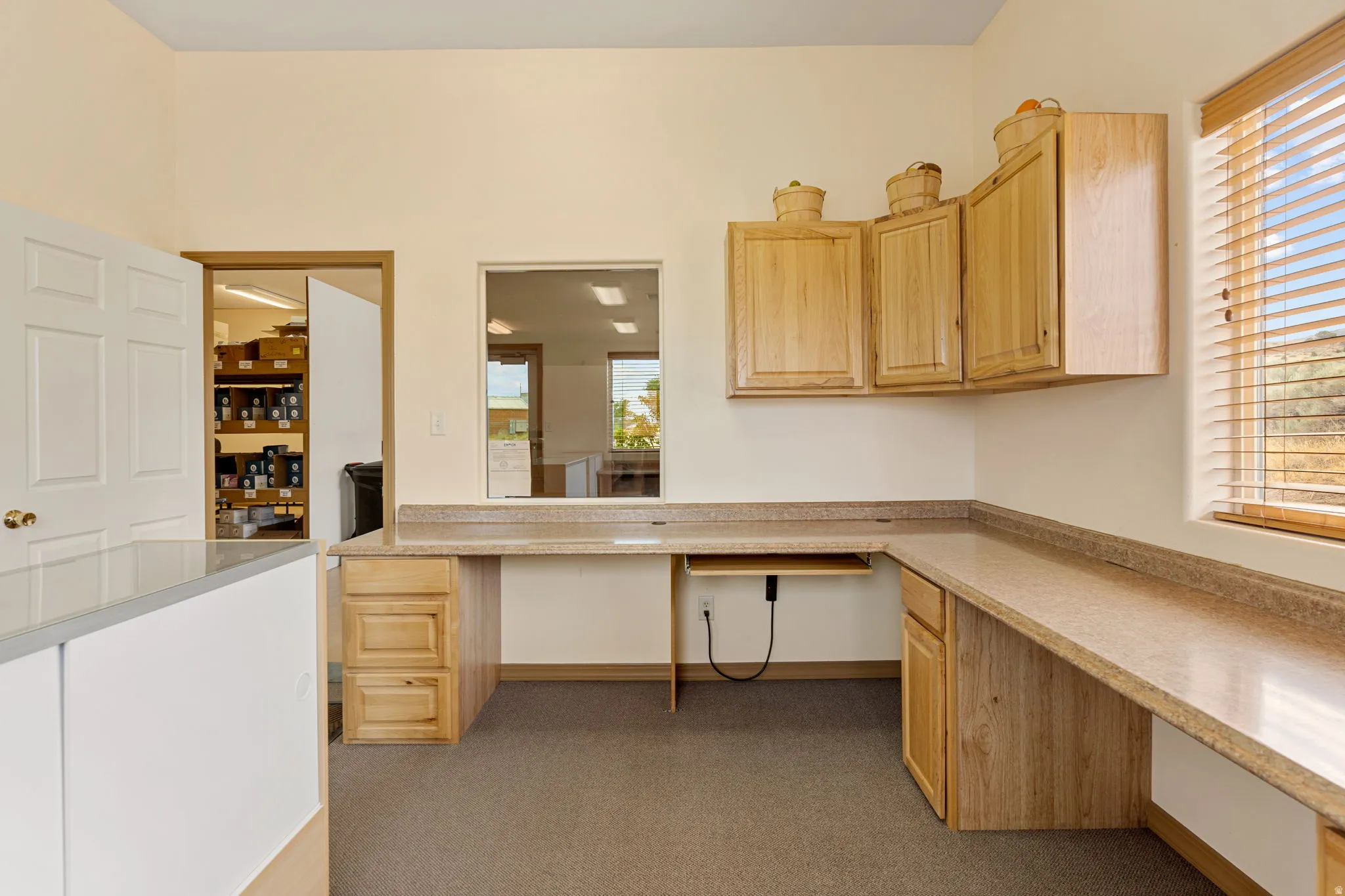 Dedicated workspace featuring extensive L-shaped countertops, ample built-in cabinetry, carpet flooring, a window with horizontal blinds, and a pass-through window
