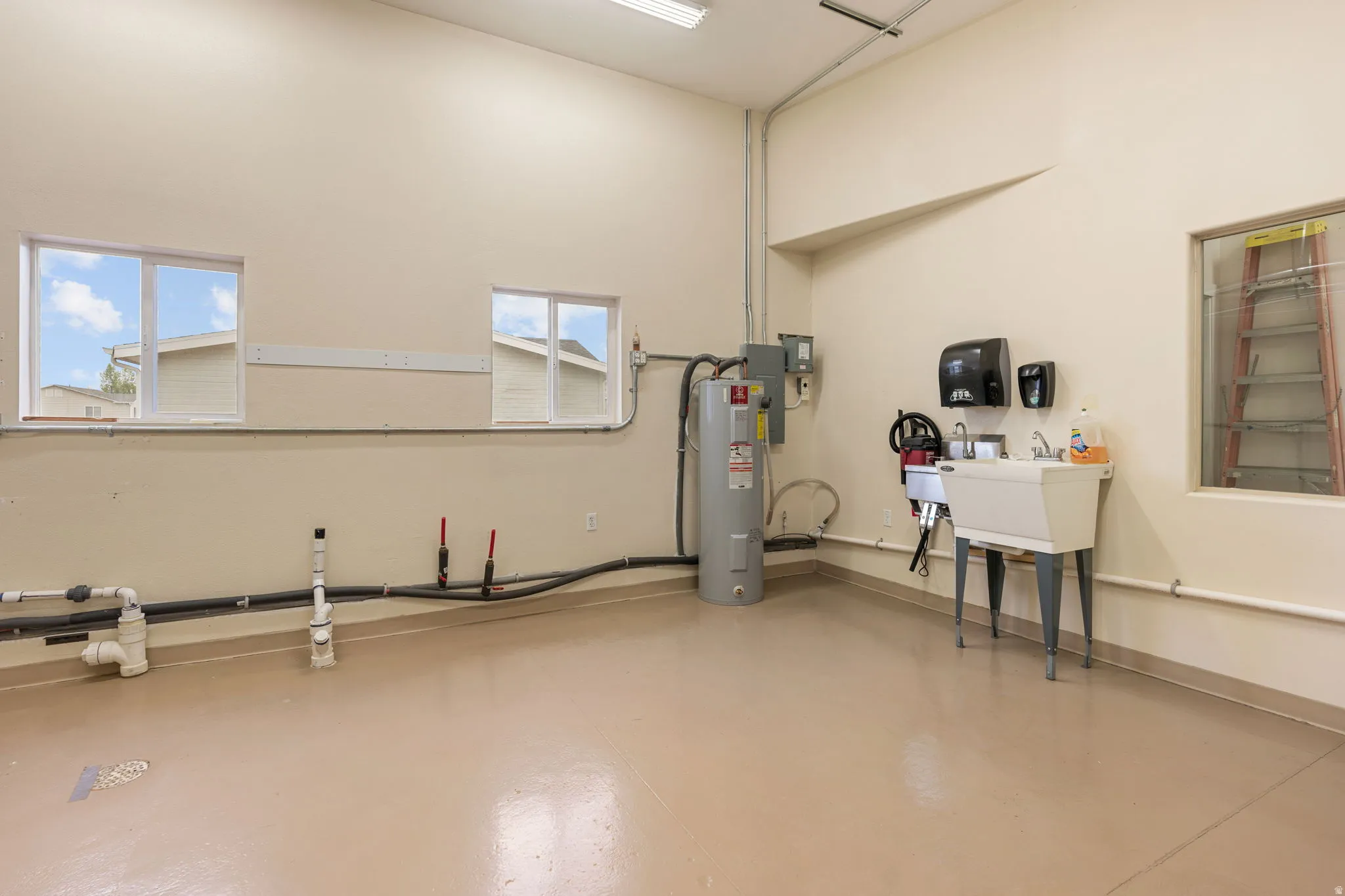 Spacious utility room featuring a utility sink, water heater, high ceilings, multiple windows, and smooth concrete flooring