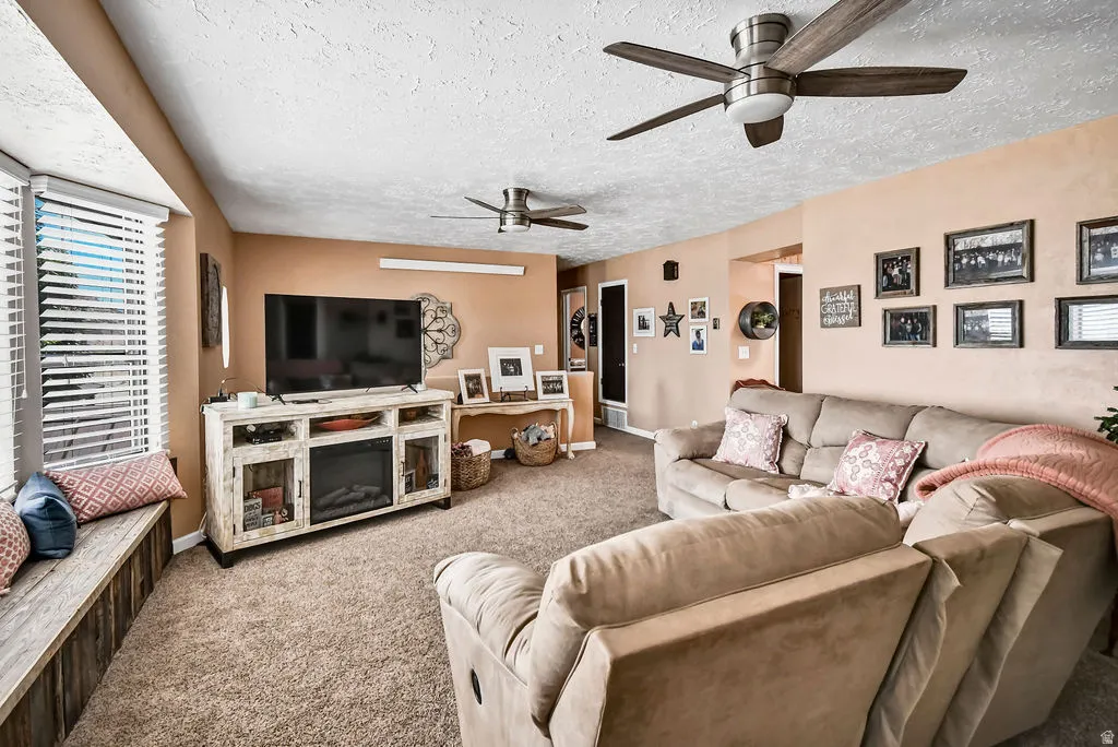 Spacious living area featuring neutral carpet flooring, a built-in window bench, a media console with an integrated electric fireplace, and two ceiling fans with wood-finish blades