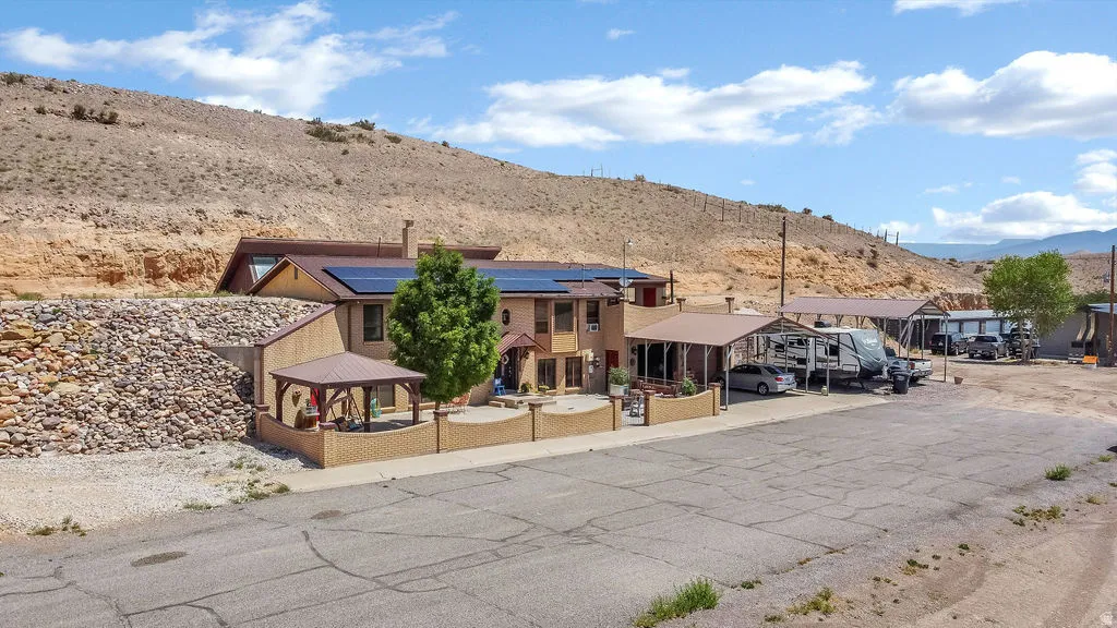 Desert landscape property featuring multiple covered parking structures, solar panels, a rock retaining wall, a paved driveway, and a brick-finish front wall with a gazebo