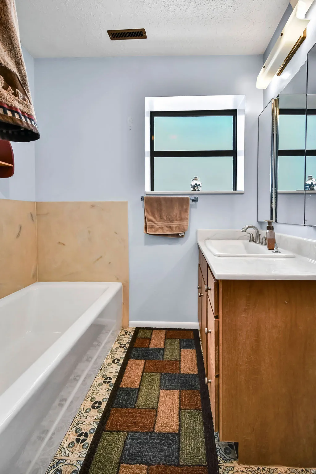 Bathroom featuring a wood-finish vanity with a white countertop and integrated sink, a white bathtub with light-toned wall tile surround, and a window with a mirrored border