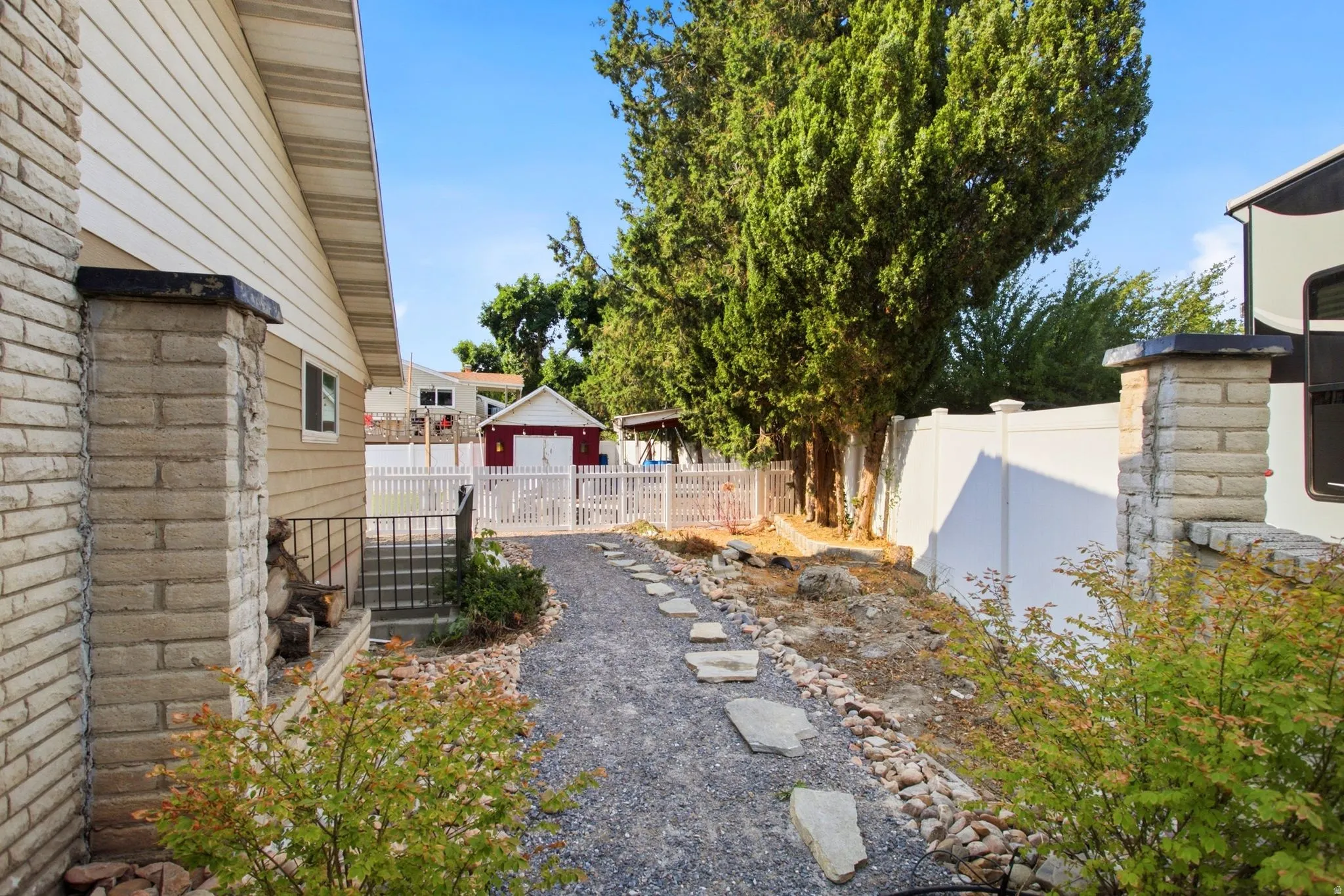 Gravel pathway with stone steppers leading to a white picket fence, complemented by a white vinyl privacy fence and established trees
