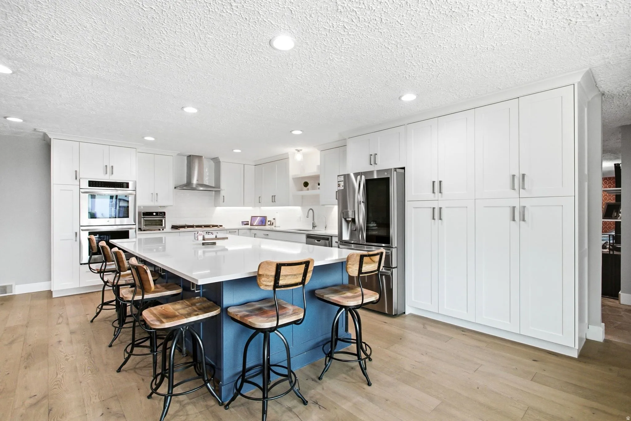 Spacious kitchen featuring an expansive center island with seating, white shaker cabinetry, stainless steel appliances, wood-finish flooring, and recessed lighting