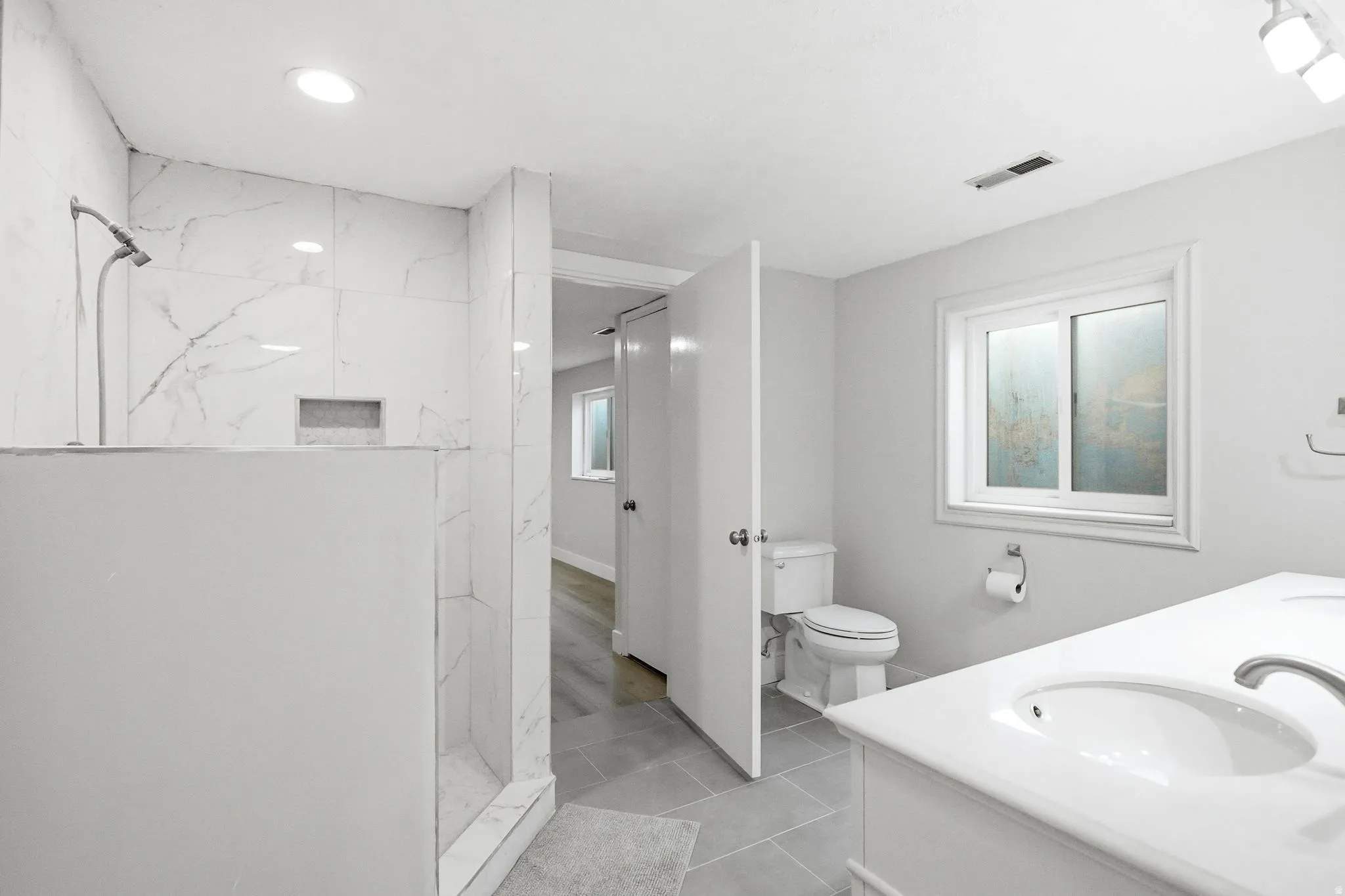 Spacious bathroom featuring a walk-in shower with large format tile surround and built-in niche, dual sink vanity with white countertop, and tiled flooring