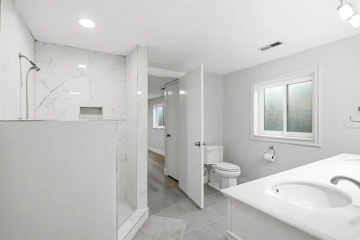 Spacious bathroom featuring a walk-in shower with large format tile surround and built-in niche, dual sink vanity with white countertop, and tiled flooring