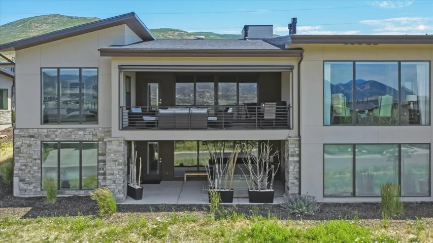 Contemporary rear exterior featuring expansive multi-pane windows, a covered upper-level balcony with metal railings, a covered lower-level patio, and natural stone veneer accents