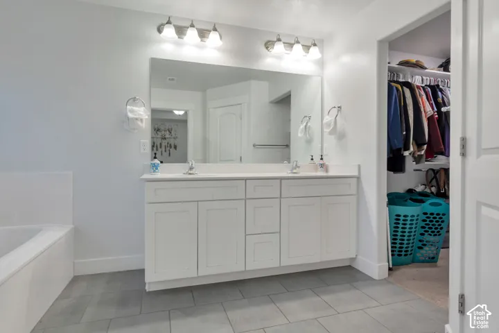 Spacious bathroom featuring a double vanity with white cabinetry, a large integrated mirror, overhead vanity lighting, a built-in bathtub, and gray tile flooring
