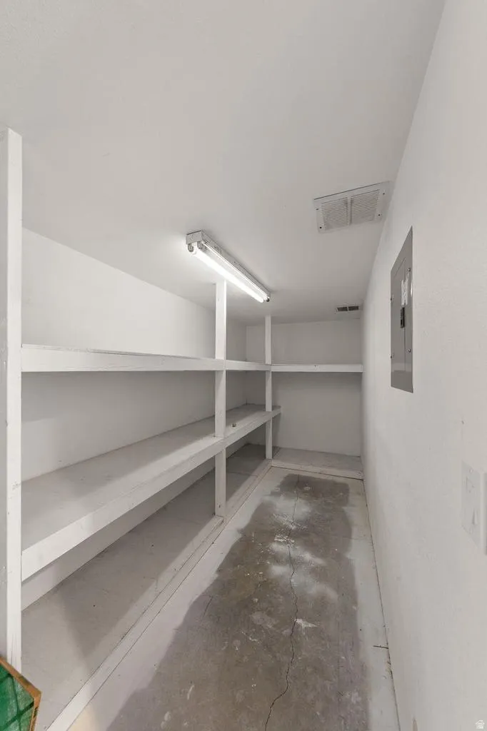 Storage area featuring built-in shelving, concrete flooring, overhead fluorescent lighting, and a wall-mounted electrical panel