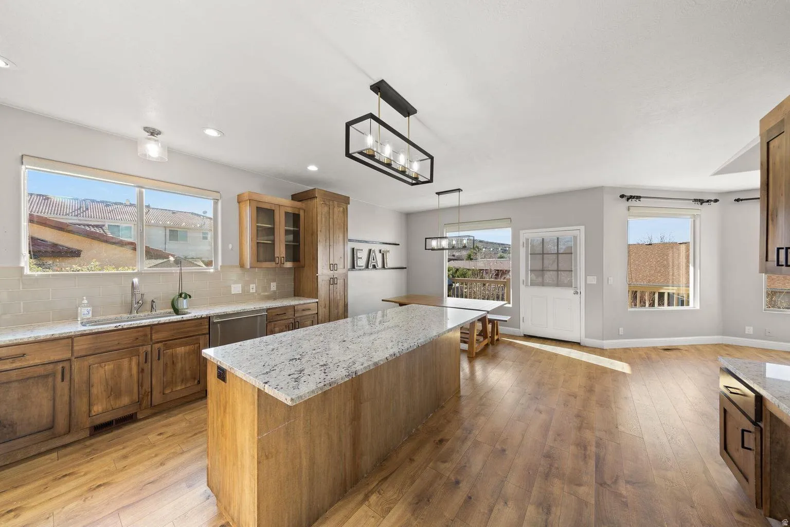 Open-concept kitchen featuring wood-finish flooring, a large island with a light-toned countertop, wood cabinetry, stainless steel appliances, and multiple windows