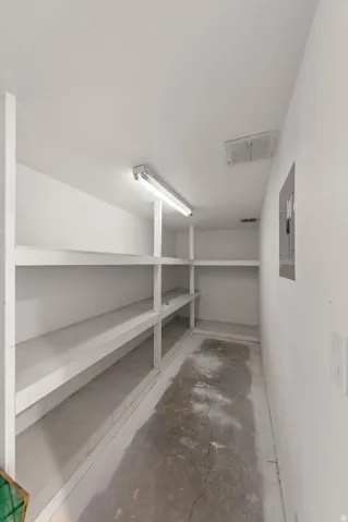 Storage area featuring built-in shelving, concrete flooring, overhead fluorescent lighting, and a wall-mounted electrical panel
