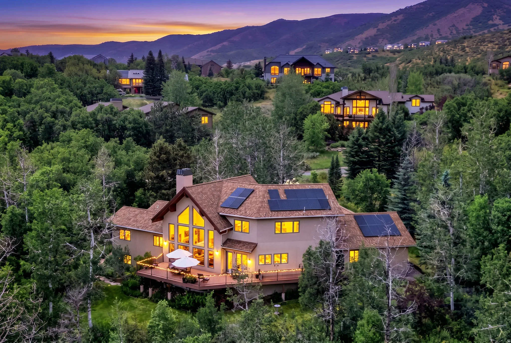 Mountain-surrounded property featuring a large deck, solar panel arrays, extensive window installations, a prominent chimney, and mature trees