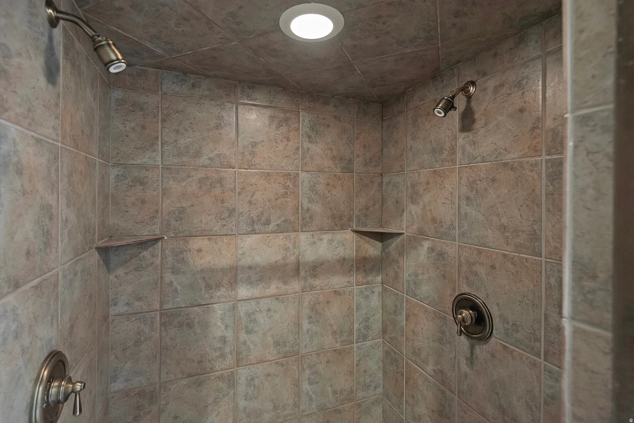 Tiled shower enclosure featuring dual shower heads with bronze-finish fixtures, recessed lighting, and integrated corner shelves
