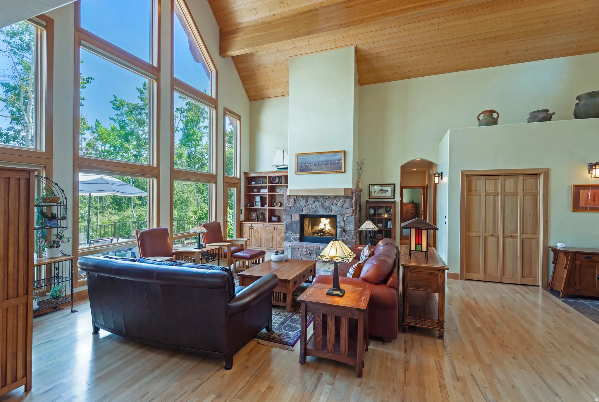 Spacious living area featuring floor-to-ceiling windows, a vaulted wood-paneled ceiling, a stone-faced fireplace, light wood-finish flooring, and natural wood trim throughout