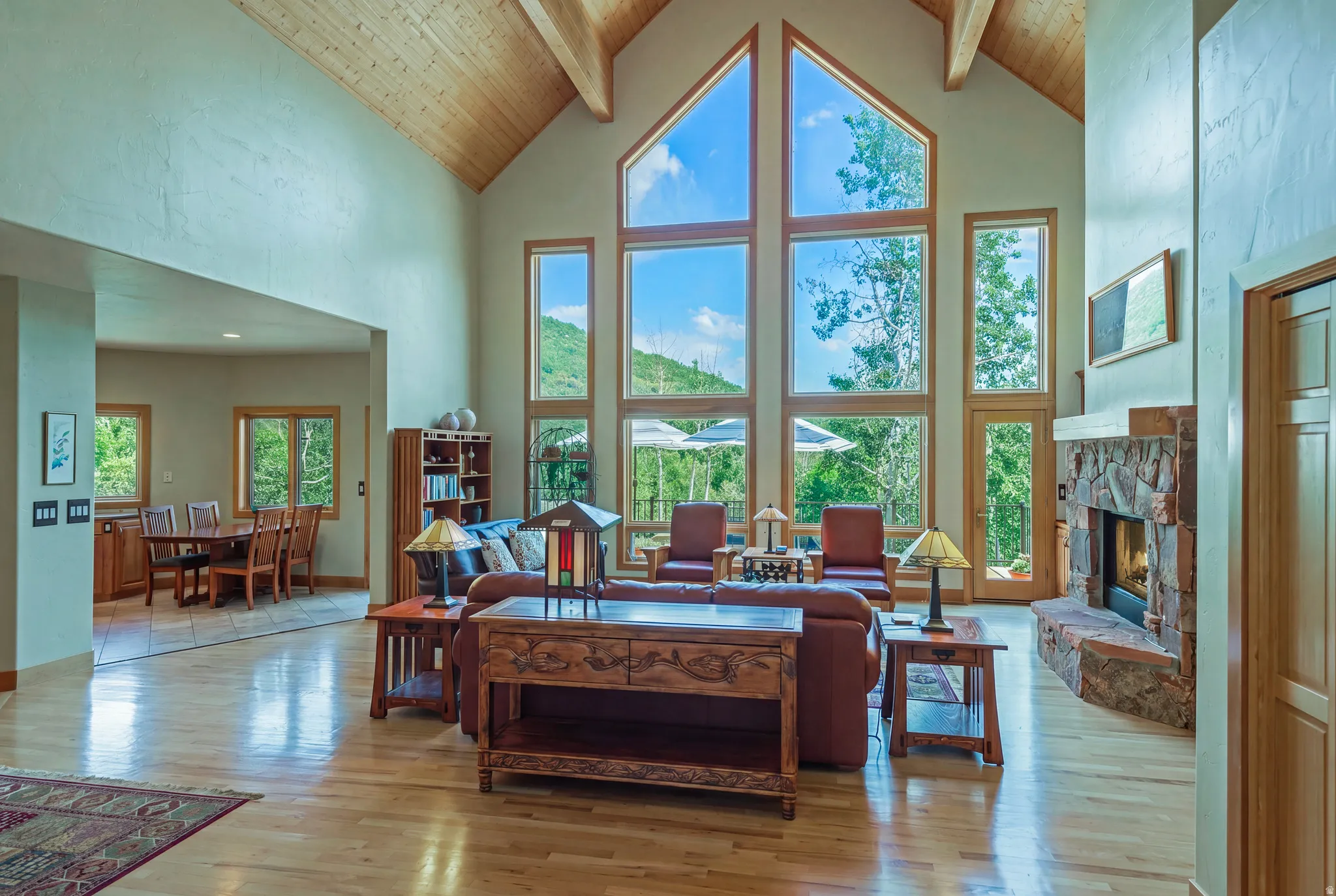 Vaulted ceilings with exposed wood beams define this spacious living area, featuring a stone-faced fireplace, light wood-finish flooring, and a dining area with tile flooring