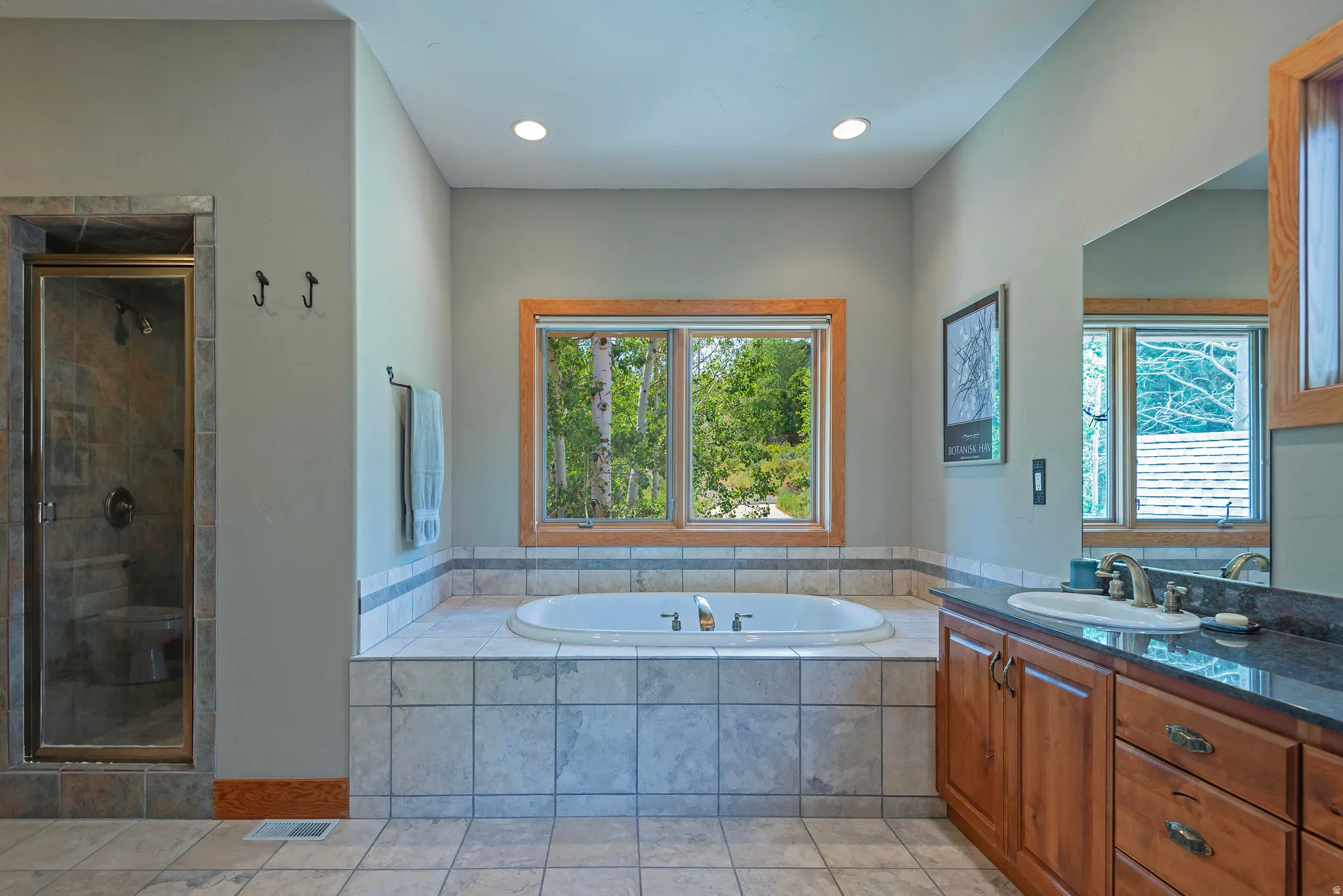 Bathroom featuring a large soaking tub with tile surround, a separate glass-enclosed shower with tile walls, a vanity with wood cabinetry and dark countertop, and a window with wood trim