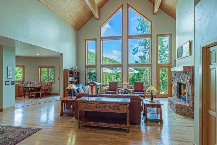 Vaulted ceilings with exposed wood beams define this spacious living area, featuring a stone-faced fireplace, light wood-finish flooring, and a dining area with tile flooring