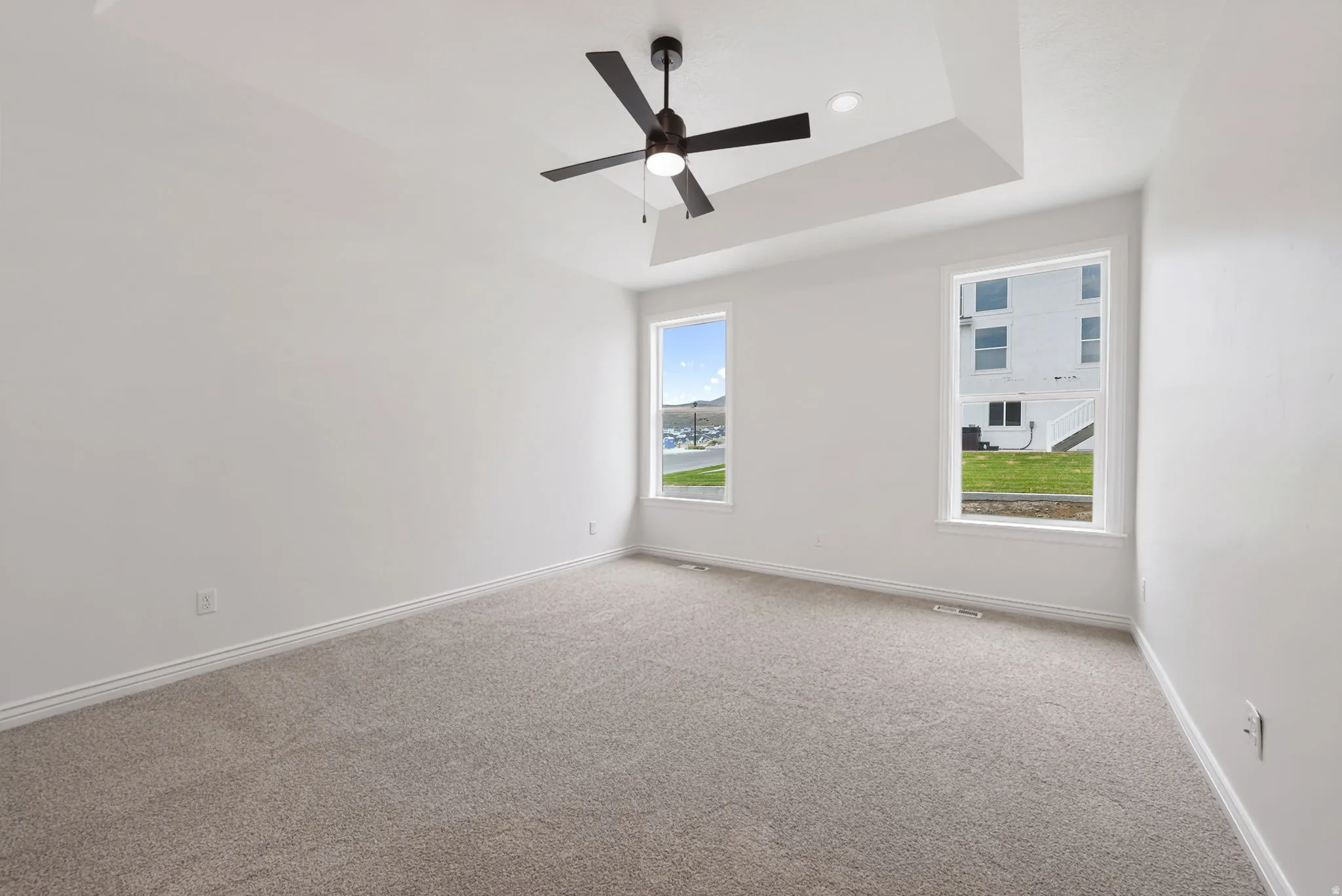 Spacious room featuring a tray ceiling with a ceiling fan, recessed lighting, two windows, and light gray carpeting