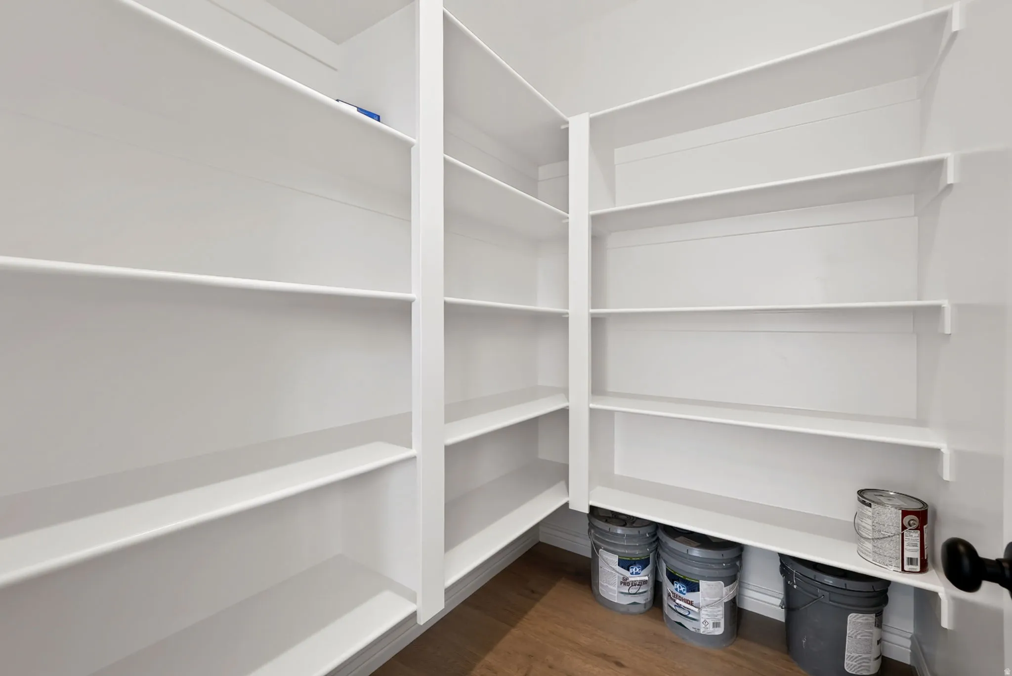 Spacious pantry featuring extensive white shelving, wood-finish flooring, and a black door handle