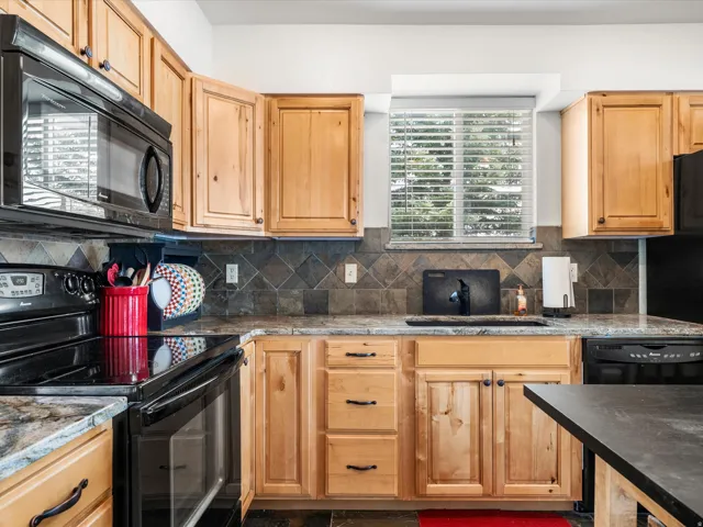 Kitchen featuring wood cabinetry, dark stone countertops, a tiled backsplash, built-in black appliances, and a window with horizontal blinds