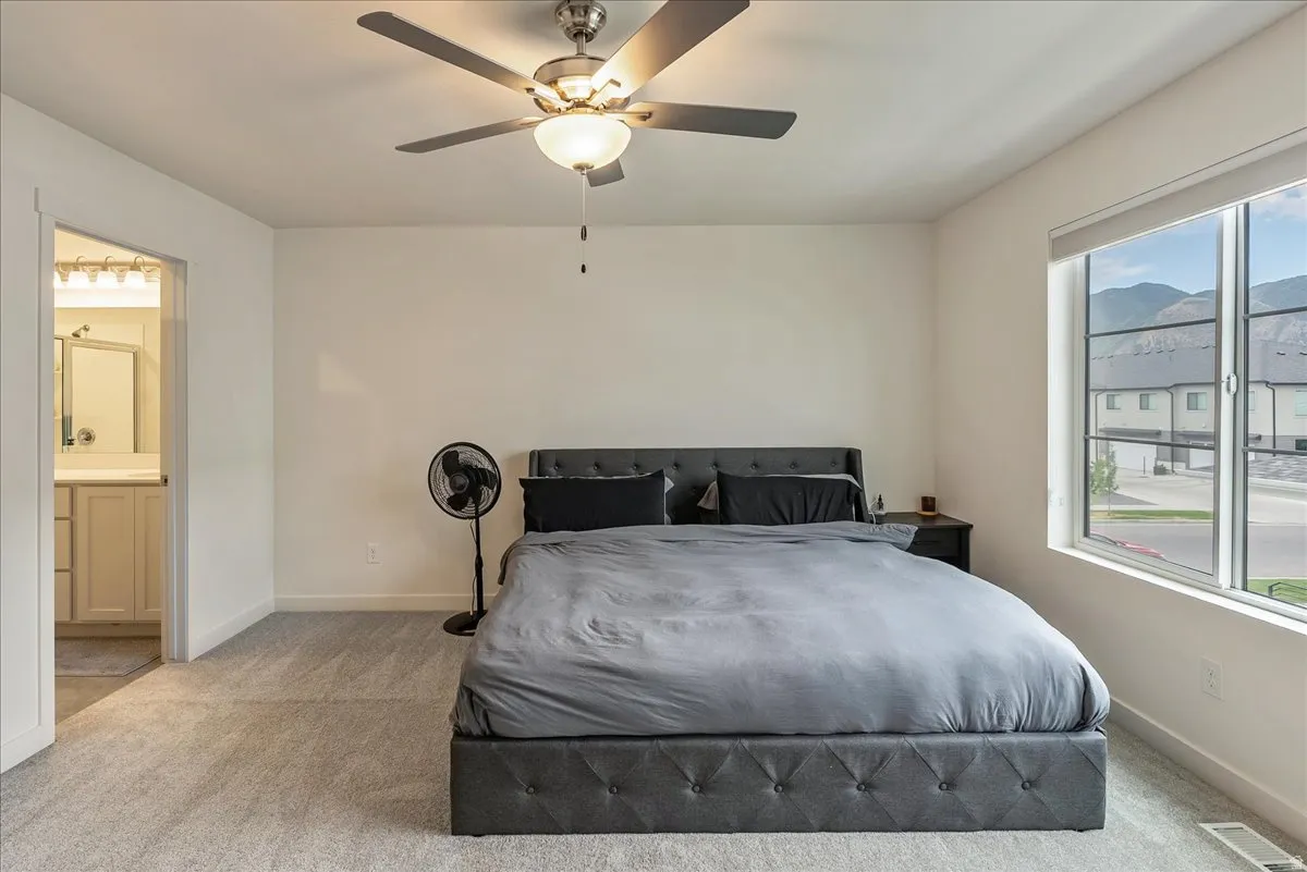 Spacious room featuring light-colored carpeting, a ceiling fan with integrated lighting, a large window with roller shades, and an open doorway revealing a bathroom vanity with a light-colored countertop