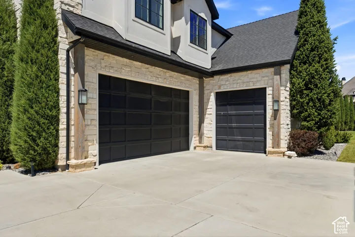 Two black sectional garage doors framed by stone siding and wood columns, complemented by exterior wall sconces, a dark shingle roof, and mature evergreen landscaping
