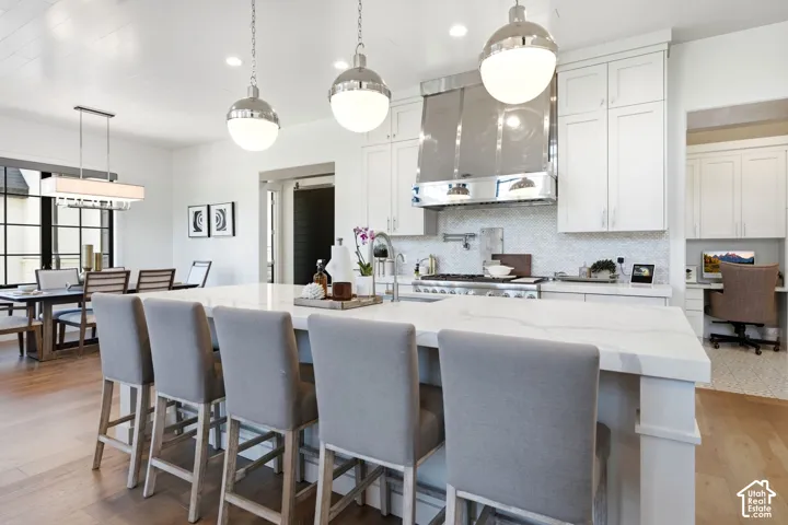 Gourmet kitchen featuring a large island with seating, pendant lighting, stainless steel range hood, white cabinetry, and a dedicated desk alcove