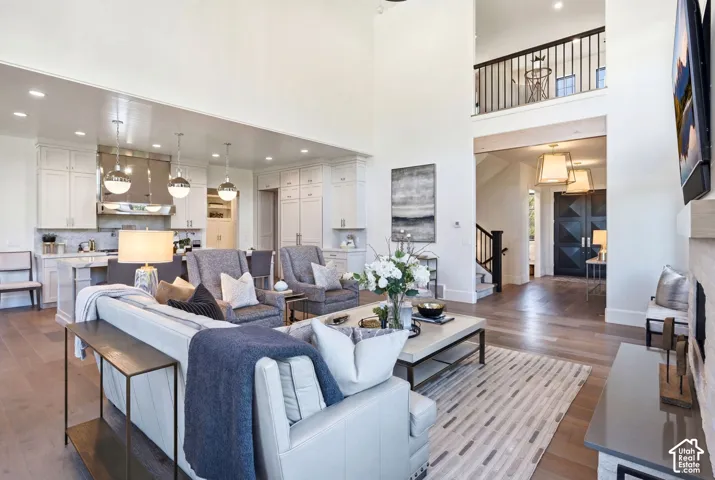 Open-concept living space featuring high ceilings, wood-finish flooring, a kitchen with white cabinetry and stainless steel appliances, a fireplace with a stone surround, and an upper-level balcony with iron railings