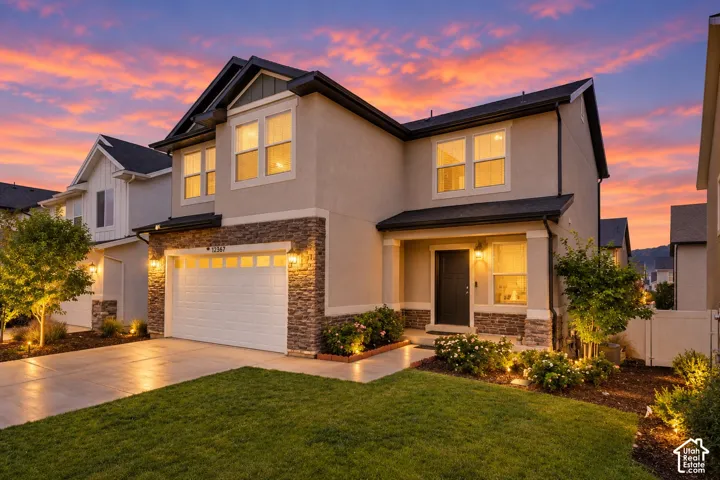Stucco exterior with stone accents, attached garage with panel door, front porch entry, well-maintained lawn, and established landscaping