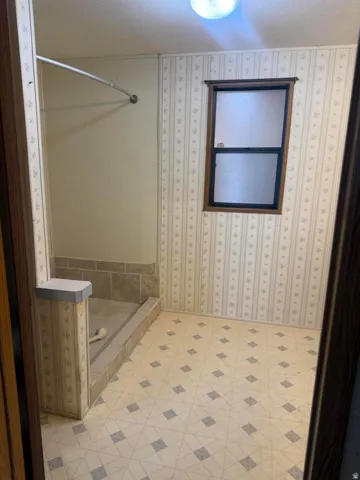 Bathroom featuring a built-in shower enclosure with a tiled base and walls, a frosted window with dark wood trim, patterned wallpaper, and geometric pattern flooring