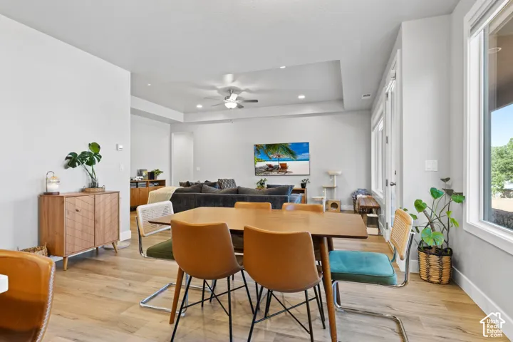 Open-concept dining and living area featuring wood-finish flooring, abundant natural light from multiple windows, a ceiling fan, and a tray ceiling