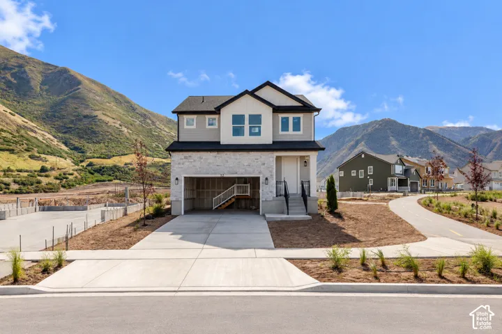 Two-story residence featuring light-toned stone veneer and horizontal siding, a covered entryway with iron railing, an attached garage, and a mountainous backdrop