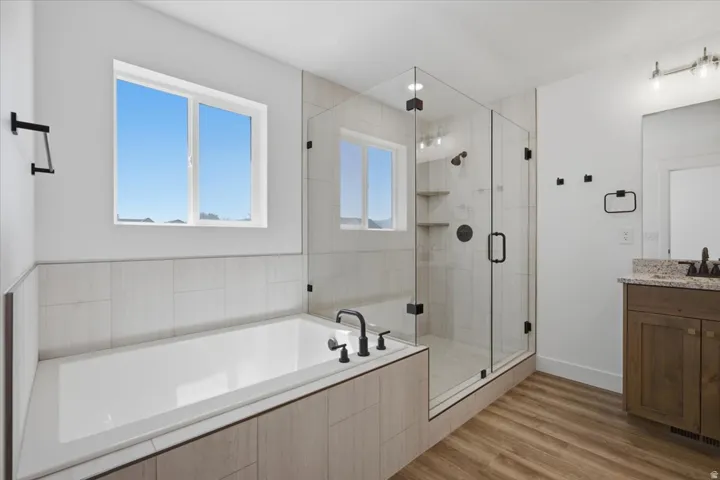 Tiled soaking tub, glass-enclosed shower with built-in shelving, wood-finish flooring, and a vanity with a stone countertop