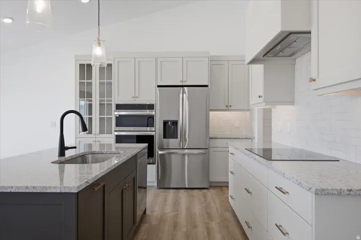 Vaulted ceiling kitchen featuring an island with an undermount sink and matte black faucet, stainless steel appliances, light gray shaker cabinetry, and wood-finish flooring