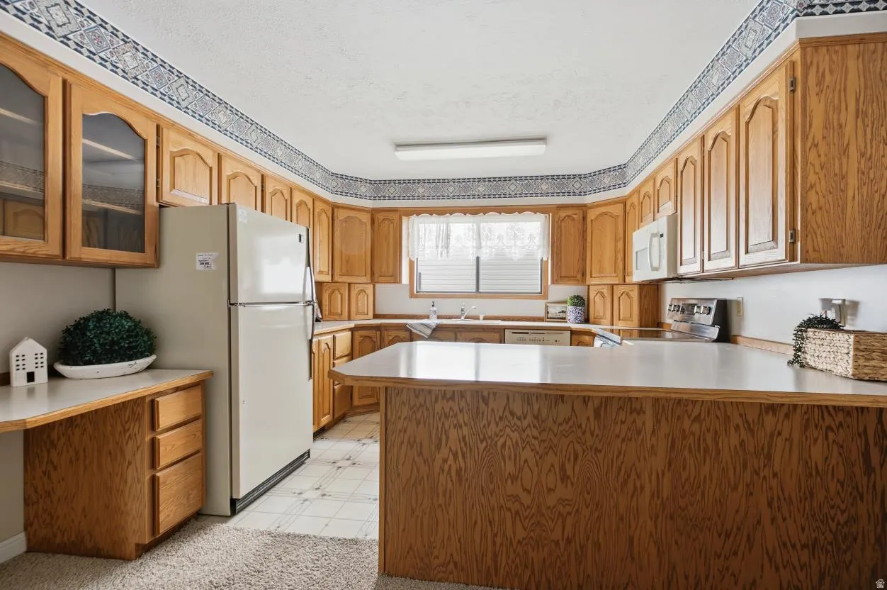 U-shaped kitchen featuring light wood cabinetry with upper glass-front cabinets, white countertops, a built-in desk area, and patterned tile flooring