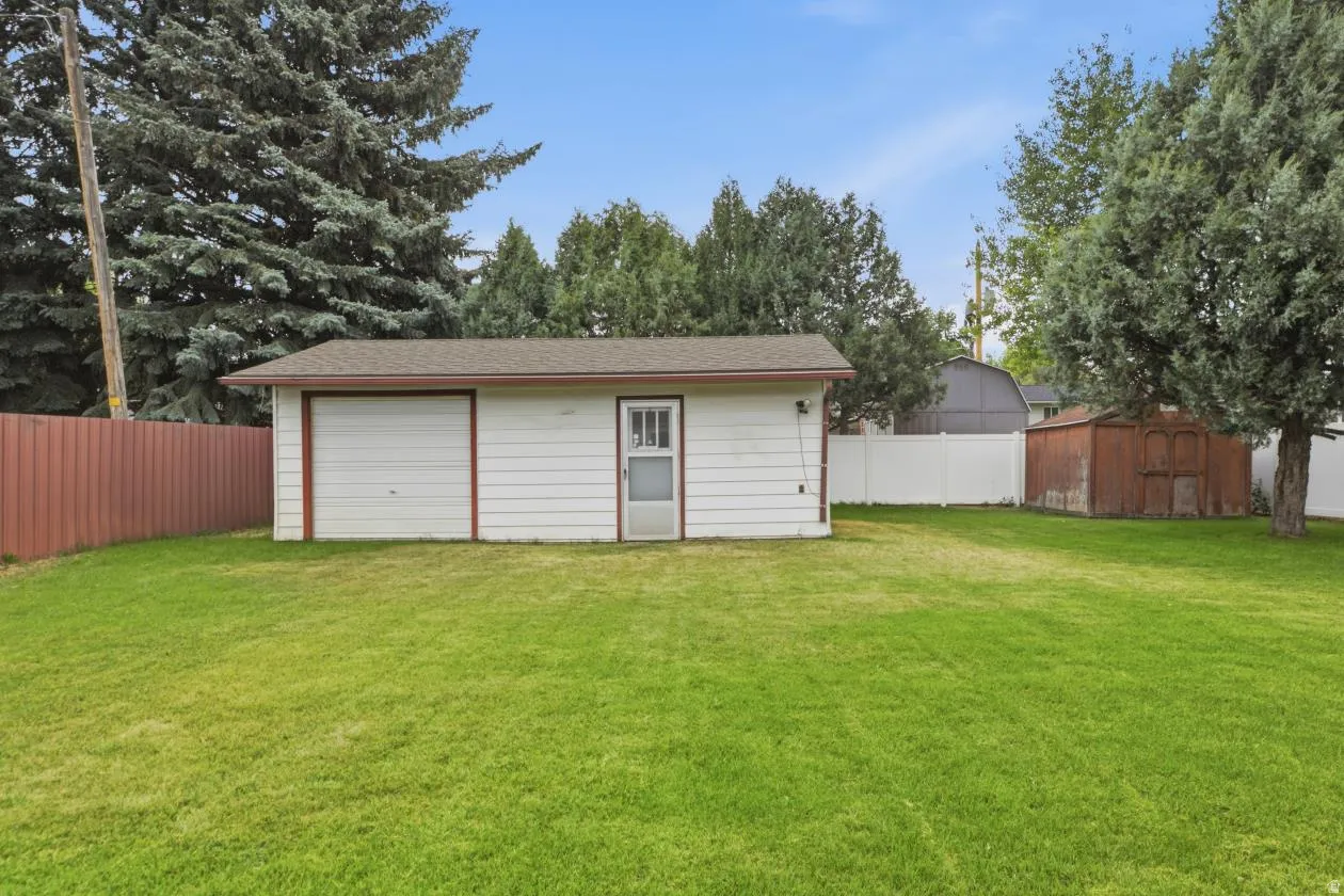 Detached garage with white siding, a single garage door, and an entry door, set within a spacious grassy area featuring mature trees and a brown privacy fence
