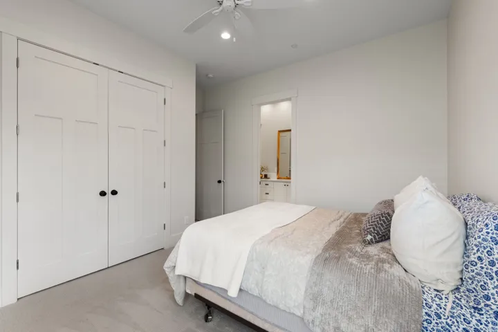 Carpeted bedroom featuring white bi-fold closet doors with black hardware, a ceiling fan with integrated lighting, and a doorway leading to a bathroom with a vanity and framed mirror